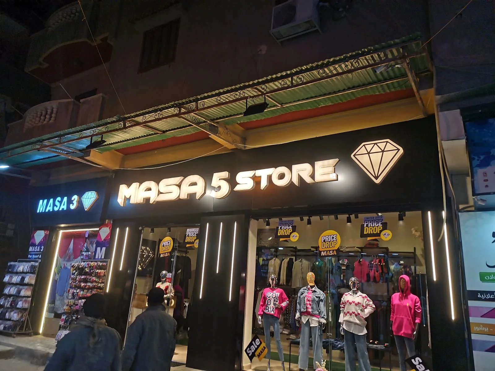 MASA 5 STORE — واجهة متجر ملابس باللون الأسود وحروف بارزة مضيئة مع خطوط إضاءة رأسية حول المدخل. من تنفيذ أبو الخير